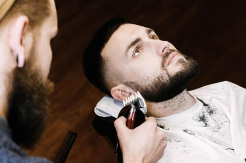 Brunette man lies with open eyes while barber cuts his beard